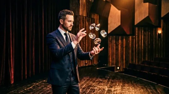 Professional magician performing on sophisticated theater stage with dramatic lighting and minimal props