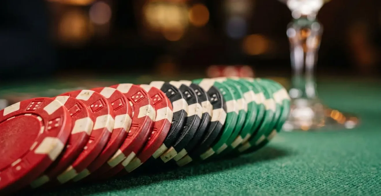 Casino gaming chips stacked on a green felt table surface with the edge of a cocktail glass in the soft-focus background.
